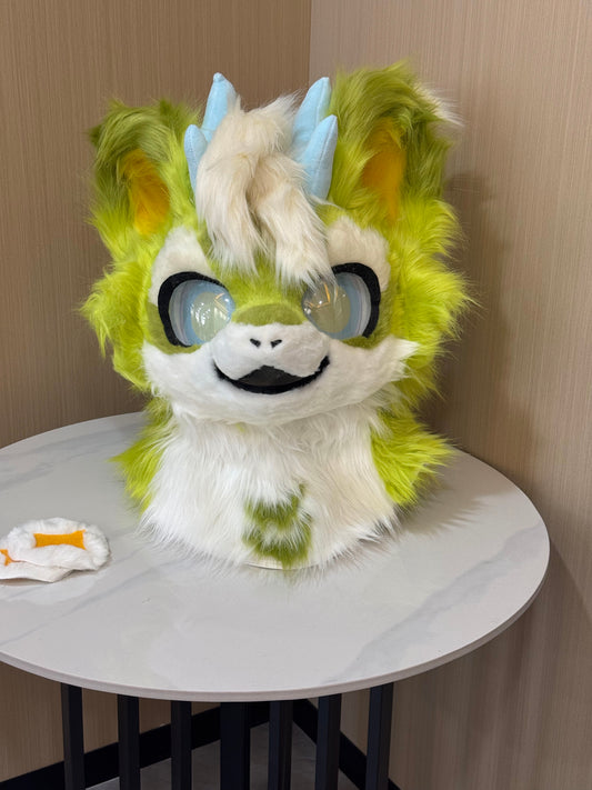 Lime green and cream feline fursuit head with blue horns and kemono-style eyes